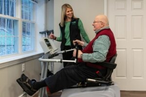 An older gentleman in a seated exercise machine smiling at the woman helping him