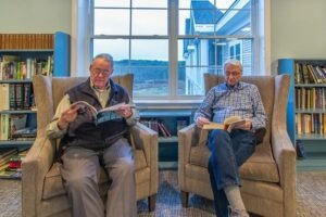 Two people sitting in lounge chairs in front of a large window reading books next to bookcases